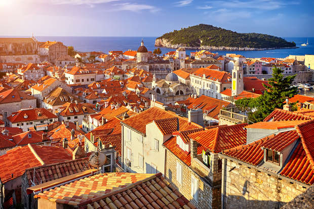 Terracotta rooftops cover a historic town, basking in sunlight. A church dome and stone buildings dominate the scene. In the background, the sea and a large, green island are visible.
