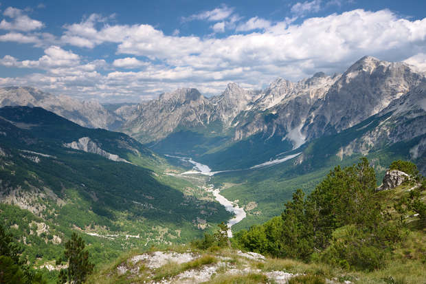 River winding through a lush, green valley, framed by towering, rugged mountains under a partly cloudy blue sky, creating a serene and majestic landscape.