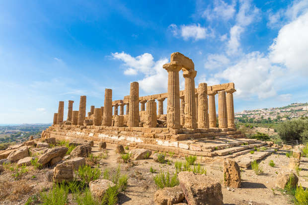Ancient stone temple ruins stand under a blue sky with scattered clouds. The temple features Doric columns amid a rocky, grassy landscape, with hills and a distant town in the background.