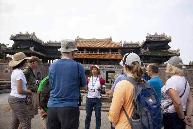 A tour guide speaks to a group of tourists in front of a large, historic, Asian-style gate with ornate rooftops, under a clear sky.