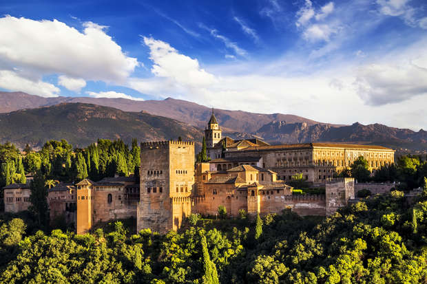 A historic stone palace with towers and intricate architecture sits atop a lush, green hill under a vibrant blue sky, with distant mountains in the background.