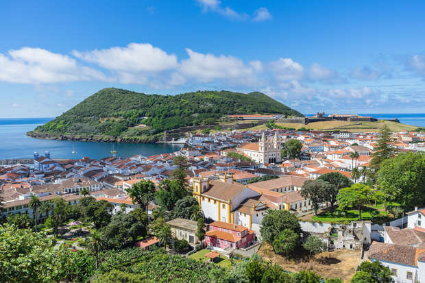 A coastal town with red-roofed buildings sits next to a harbor, surrounded by lush green hills under a partly cloudy sky, creating a picturesque landscape.