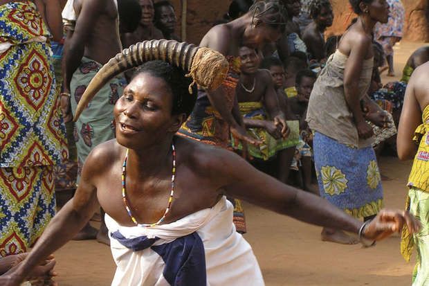 A person adorned with a large horn dances energetically, surrounded by others in colorful attire, in an outdoor cultural gathering with earthy tones.