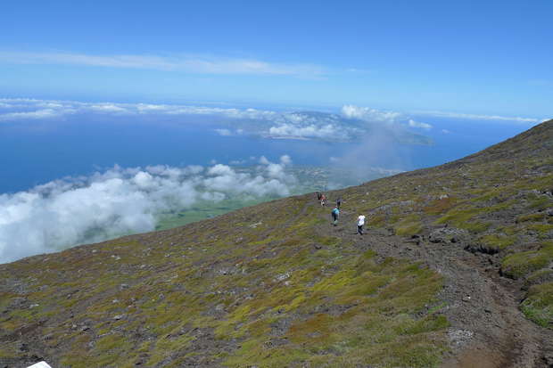 Hikers walk along a narrow trail on a sloped, grassy mountainside. Below, a vast landscape stretches out, with clouds and the ocean visible under a clear blue sky.