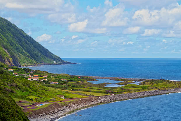 Coastal landscape with rolling hills and scattered houses stretches along a rocky shoreline. The ocean extends to the horizon, beneath a sky dotted with fluffy clouds.