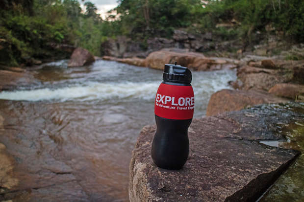 A black water bottle with a red label reading "EXPLORE First Adventure Travel Experts" rests on a rock beside a flowing river, surrounded by lush greenery.