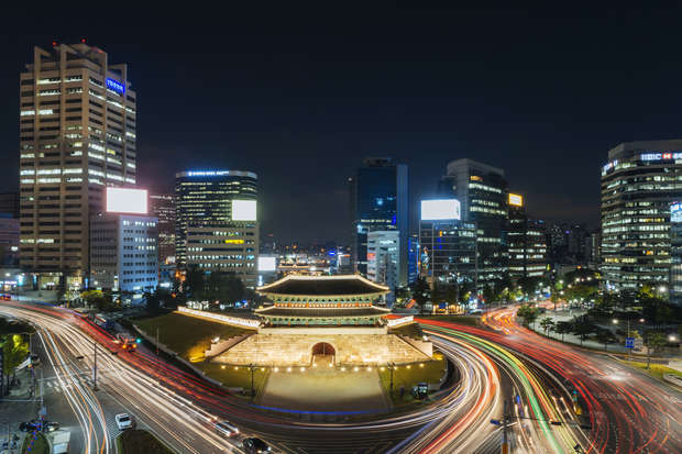 Historic gate illuminated amidst surrounding skyscrapers, with light trails of passing vehicles creating a dynamic pattern, in a bustling urban cityscape at night.