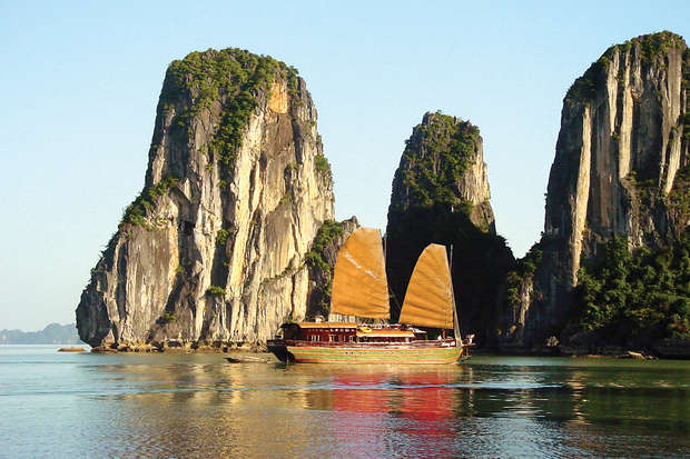 A traditional wooden boat with tan sails glides across tranquil water surrounded by towering limestone karsts, under a clear blue sky in a picturesque natural seascape.