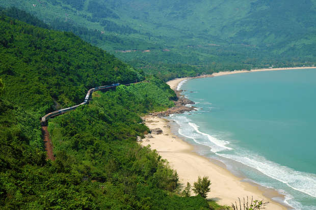 A train travels along a curving track through lush, green hills beside a sandy beach with gentle ocean waves, under a clear sky.