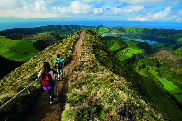 Hikers walk along a narrow dirt path atop a lush, green ridge, surrounded by rolling hills and lakes under a partly cloudy sky.