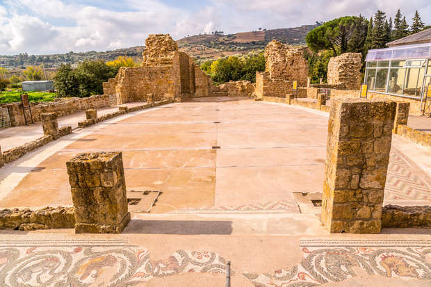 Ancient stone ruins form semi-circular structure with remnants of intricate mosaic flooring. Surrounded by lush greenery and distant hills, under a partly cloudy sky. Nearby, a modern glass enclosure.