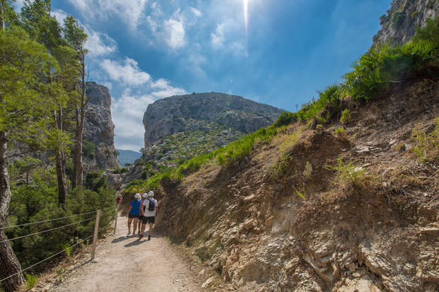 Hikers walk along a dirt path between rocky hills, surrounded by green trees and shrubs under a bright, sunny sky.