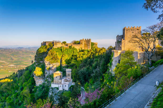 A medieval fortress sits atop a rocky, tree-covered hill, overlooking a vast landscape. Sunlight illuminates the stone walls, creating a striking contrast with the lush greenery below.