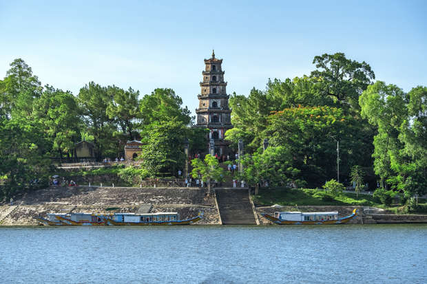 A tall, multi-tiered pagoda stands amidst lush greenery, with visitors walking the stone steps leading to it; two moored boats float on the river in the foreground.