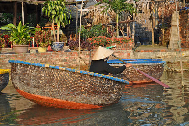 A person paddles a round bamboo basket boat on calm water, surrounded by lush potted plants and rustic wooden structures in a traditional setting.