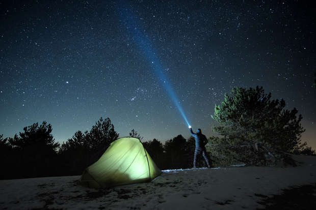 A person shines a flashlight beam toward the starry sky next to a glowing tent, surrounded by snow and trees in a nighttime forest setting.