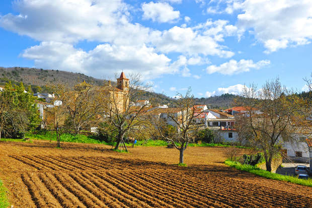A plowed field lies in the foreground with rows of soil. In the background, a village with a prominent church tower is surrounded by trees and hills under a partly cloudy sky.