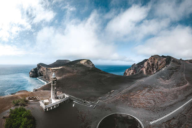 A lighthouse stands on a rocky, volcanic coastline beside the ocean. Surrounding barren hills and cliffs create a dramatic, isolated landscape under a partly cloudy sky.