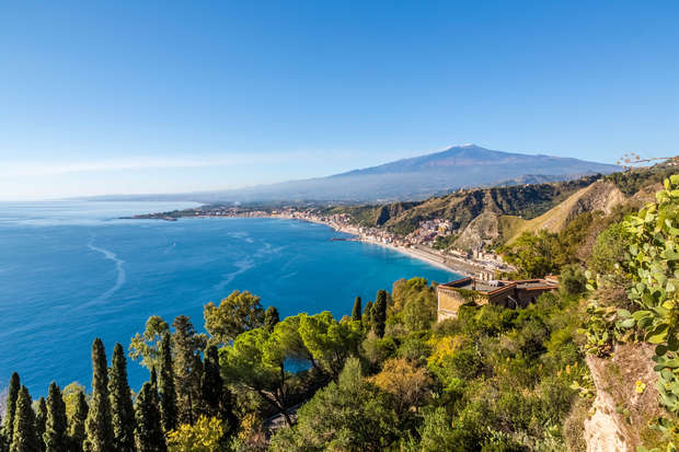 Coastal town stretches along a clear blue bay; lush greenery and steep cliffs surround, with a distant volcano towering under a clear sky. Trees and buildings dot the landscape.