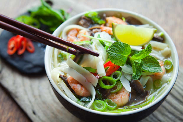 A bowl of noodle soup with shrimp is held by chopsticks, garnished with lime and mint, surrounded by sliced chili and herbs on a wooden table surface.