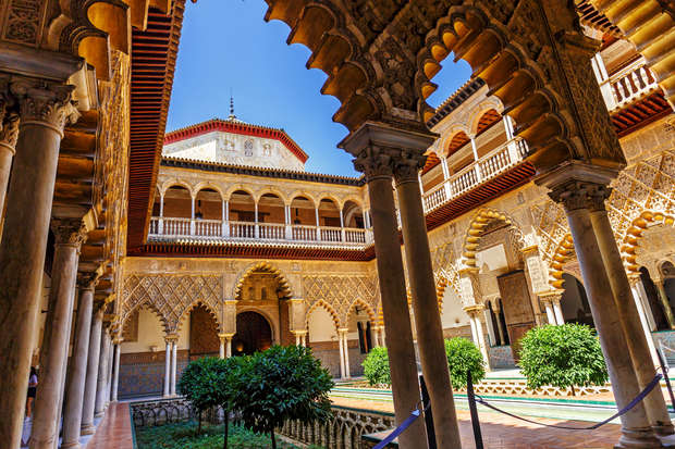 Ornate arches frame a courtyard with a rectangular pool, surrounded by lush greenery and intricate Moorish architecture. Sunlight illuminates detailed carvings and geometric patterns under a clear blue sky.