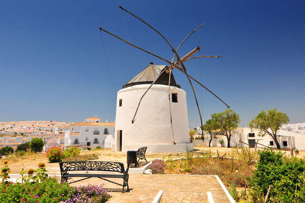 A white windmill with extended sails stands in a garden with colorful flowers and a bench. It overlooks a village of white buildings under a clear blue sky.