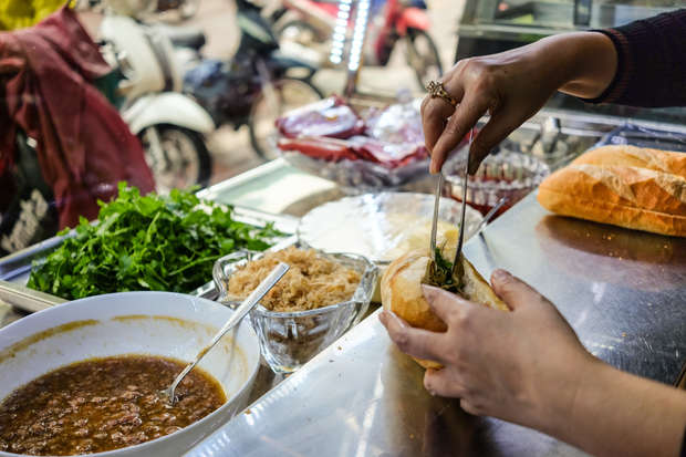 Hands prepare a sandwich by adding ingredients with tongs, surrounded by fresh herbs, sauce, and a loaf on a metal countertop in a bustling street food setting.