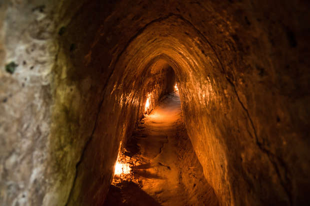 A narrow, curved earthen tunnel illuminated by small, scattered lights, recedes into darkness. The walls and floor are rough, creating a natural, cave-like atmosphere.