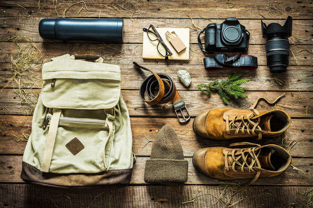 A neatly arranged flat lay of outdoor gear on wooden boards features a beige backpack, leather boots, camera with lenses, glasses, notebook, belt, thermos, beanie, and greenery, suggesting adventure preparation.
