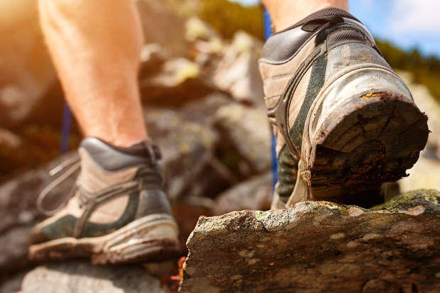 Worn hiking boots climb over rugged rocks, set against a blurred natural landscape with sunlight highlighting the surroundings.