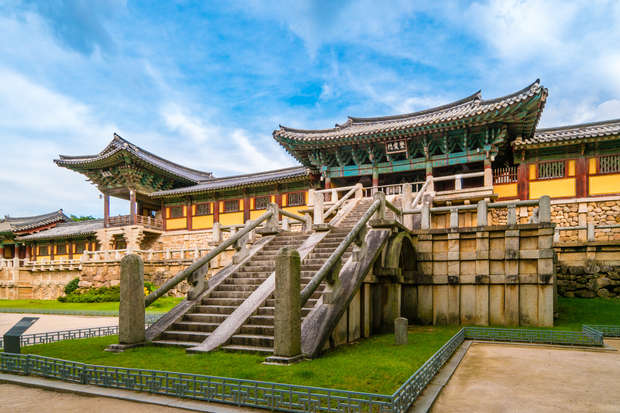 A traditional Korean temple with wooden architecture and intricate roof designs stands prominently, featuring a large stone staircase leading up to it, surrounded by manicured grass and a serene atmosphere.