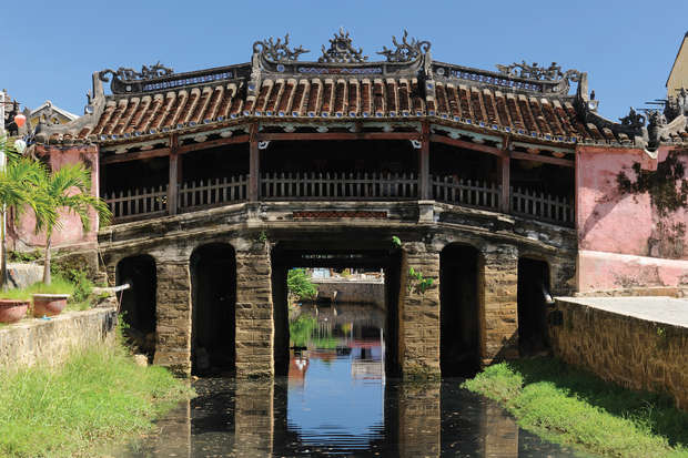 A traditional covered bridge with ornate decorations spans over a calm waterway, surrounded by pink walls and vibrant greenery under a clear blue sky.