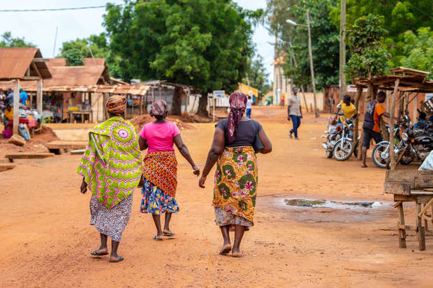 Three people walk along a dusty, orange road in a village, surrounded by small wooden structures, motorcycles, and lush green trees.