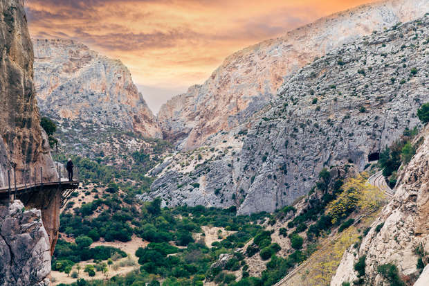 A narrow footpath clings to a steep cliffside, with a person walking along it. The rocky canyon landscape features a lush green valley below and a vibrant, colorful sunset overhead.