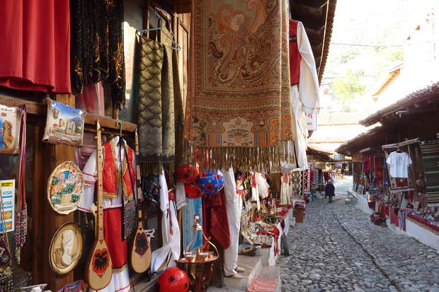 Street market displays colorful textiles and crafts on a cobblestone path, with hanging rugs and traditional clothing. Various souvenirs and art pieces are showcased outside wooden shops, creating a vibrant atmosphere.