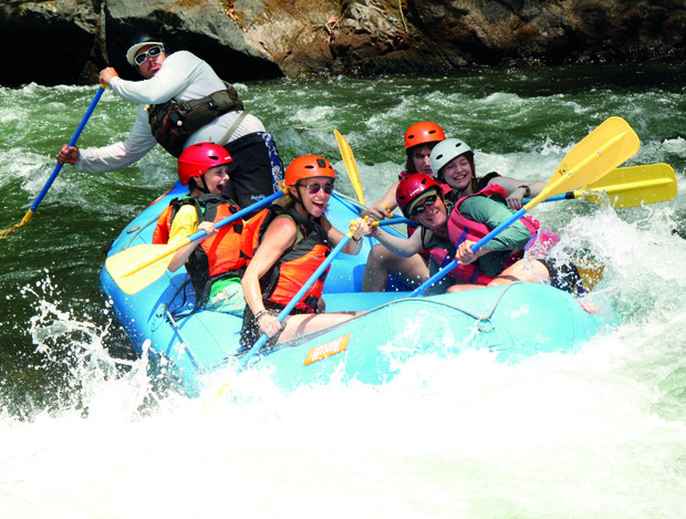 A group of people is rafting down a river, wearing helmets and life vests, paddling energetically through white-water rapids in a blue inflatable raft. Rocks line the riverbank.