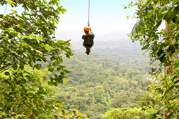 A person wearing a helmet is zip-lining, soaring above a lush, green forest canopy, surrounded by dense foliage under a clear sky.