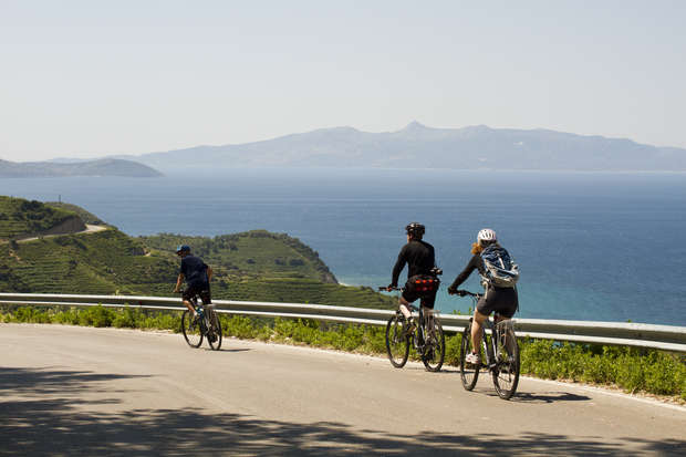 Three cyclists ride along a coastal road, overlooking rolling green hills and vast ocean with distant mountains under a clear sky.