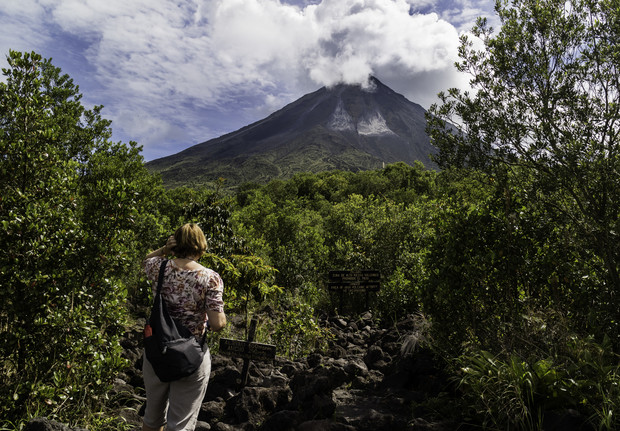 A person walks along a rocky path surrounded by lush greenery, looking at a smoking volcano in the background. Signs are visible among the vegetation.