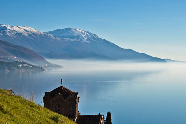 A church with a cross on its roof stands on a hillside, overlooking a calm lake with mist, against a backdrop of snow-capped mountains under a clear blue sky.