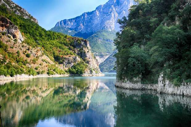 A calm river reflects rugged, forested mountains under a clear blue sky, flanked by steep, rocky cliffs and lush greenery, creating a peaceful, natural landscape.