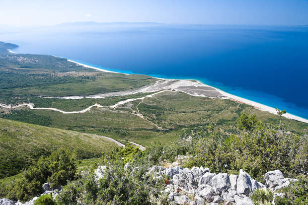 A rocky cliff overlooks a vast, green landscape leading to a bright sandy beach with deep blue sea beyond, under a clear sky. Curving paths traverse the greenery.