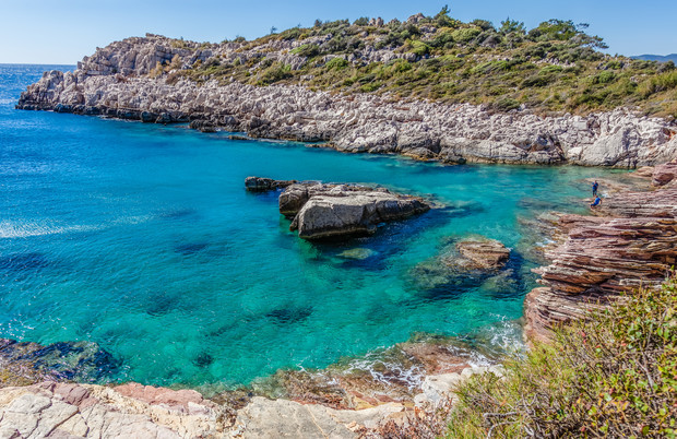 Clear turquoise water surrounds rocky cliffs, with a person standing on a cliff edge. The coastline features rugged terrain with sparse greenery, under a bright, sunny sky.