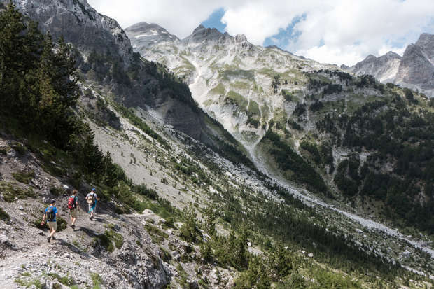 Hikers trek along a rocky mountain trail, surrounded by steep, forested slopes and rugged peaks under a partly cloudy sky, creating a dramatic alpine landscape.