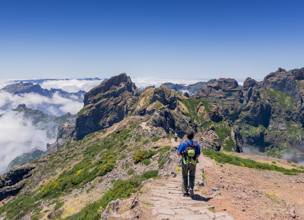 A hiker walks down a stone path surrounded by lush green mountains and distant, low clouds under a clear blue sky.