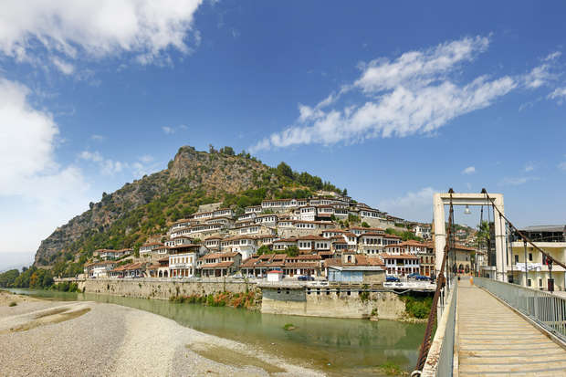 White stone houses with red roofs cascade down a steep hillside, overlooking a tranquil river. A suspension bridge crosses the river, connecting to the scenic town under a clear blue sky.
