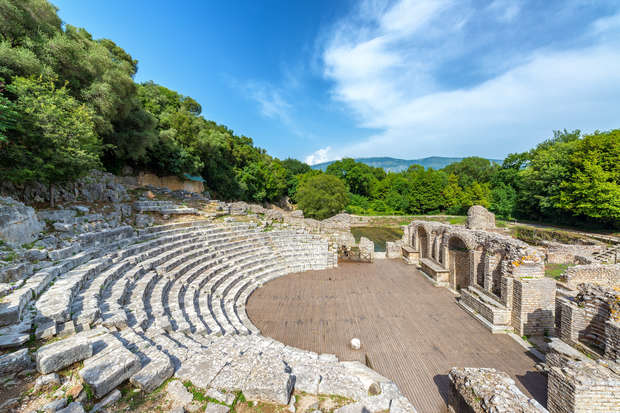 An ancient stone amphitheater with semi-circular rows of seating remains empty under a clear blue sky, surrounded by trees and distant hills, creating a serene, historical setting.