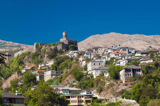 A hillside village with stone houses climbs toward a castle atop the hill, set against a backdrop of arid mountains and a clear blue sky.