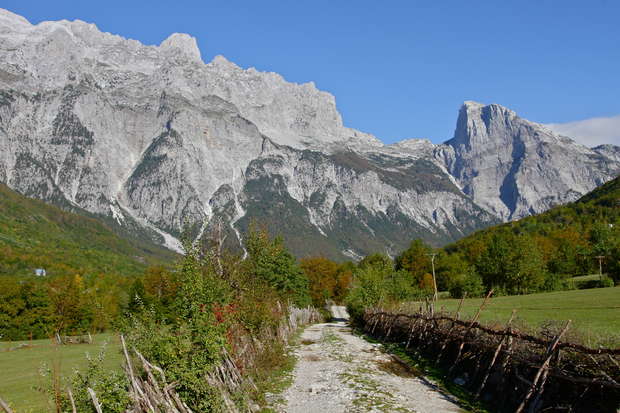 A rugged dirt path winds through a lush valley, bordered by rustic wooden fences, with towering, jagged mountains under a clear blue sky in the background.