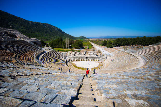 Ancient stone amphitheater with tiered seating; visitors walking down steps; surrounded by lush green landscape and distant hills under a clear blue sky.